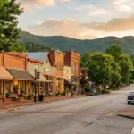 Historic brick storefronts and shade trees line a quiet main street in a small North Georgia mountain town, with a forested ridgeline visible in the late-afternoon light
